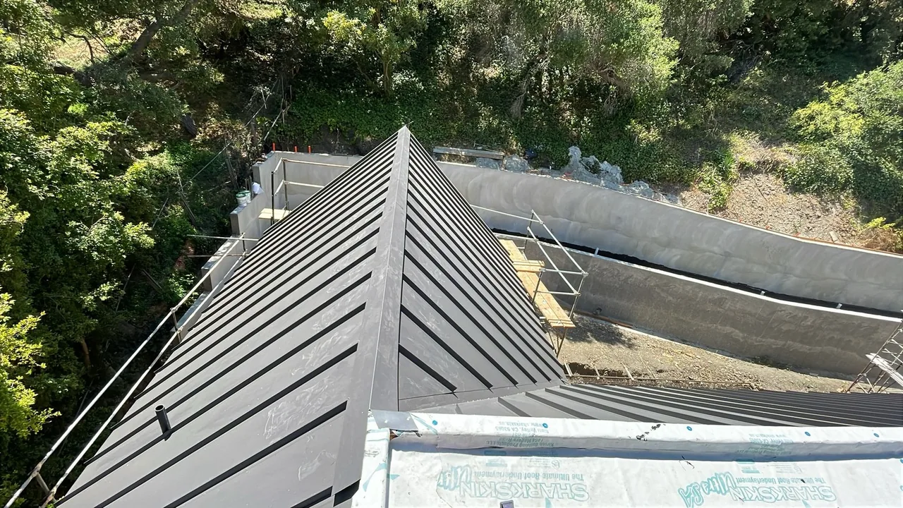 Gray standing-seam metal roof panels meeting at a hip, with white underlayment still exposed on the lower slope
