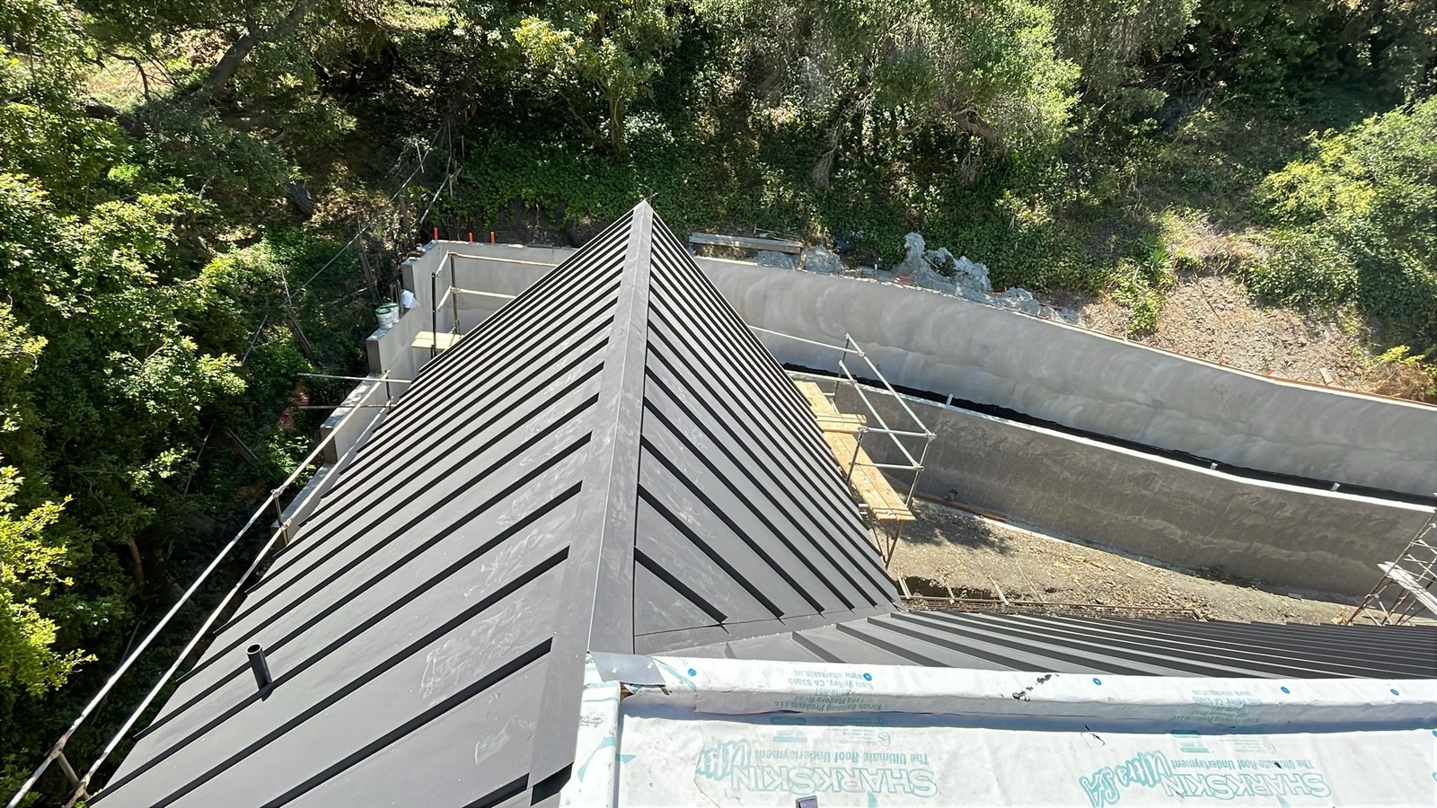 Dark gray standing seam metal roof panels installed over white underlayment on a hillside home.