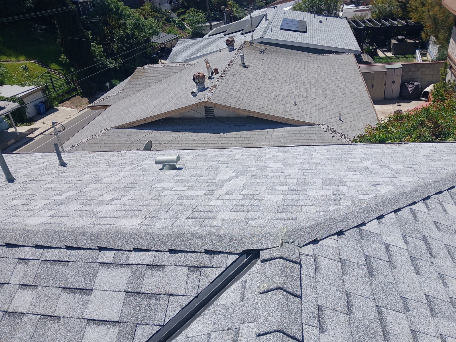 Gray asphalt shingle roof slopes with pipe vents and turbine vents above a hillside street