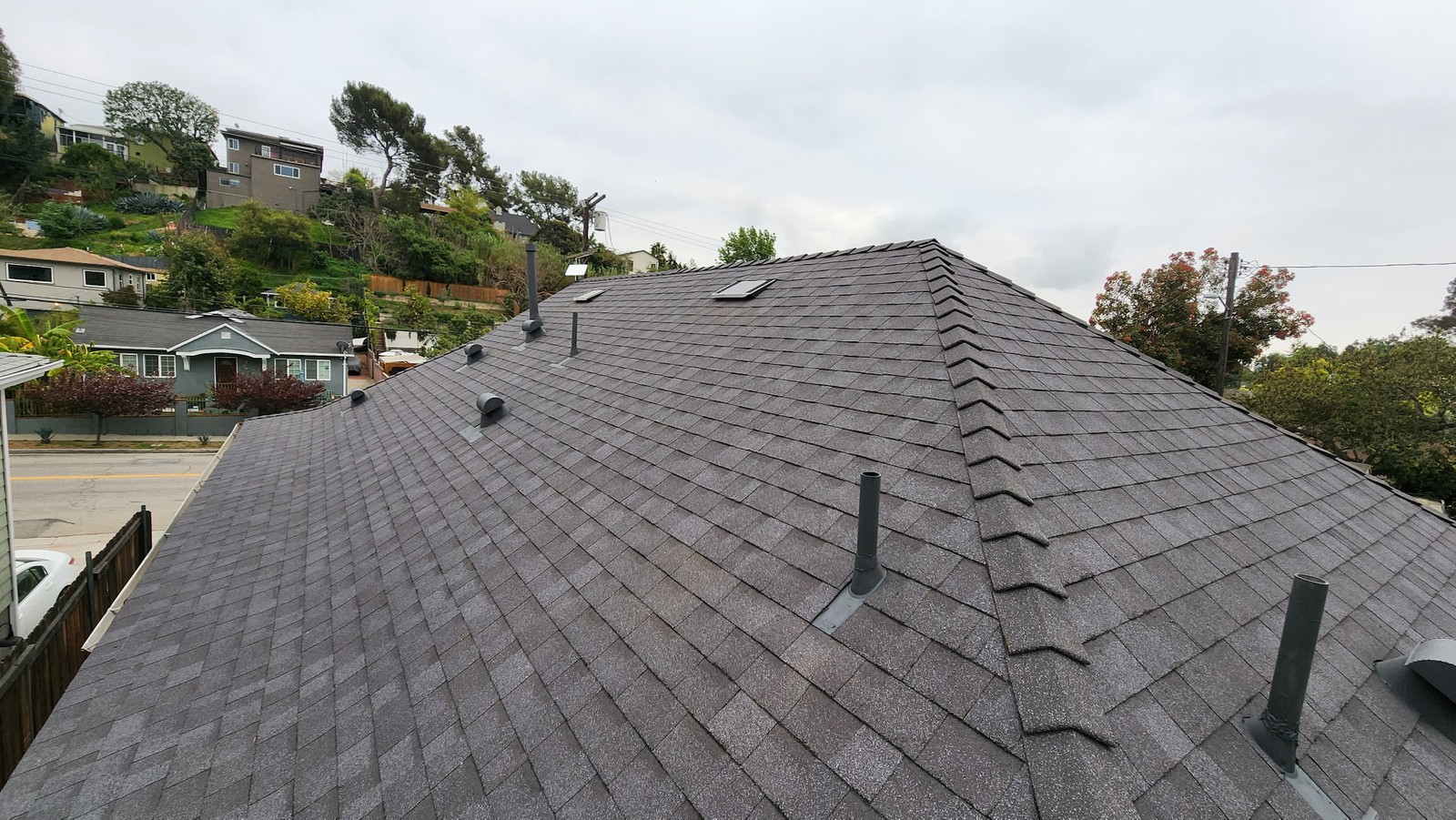 Dark gray asphalt shingle roof with ridge caps and several vent pipes, with houses on a hill behind