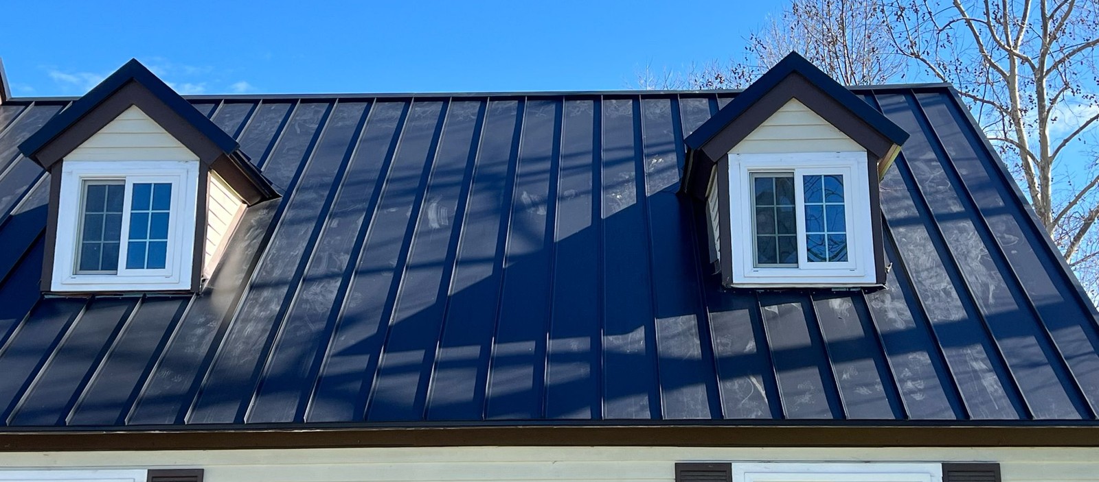 Dark standing-seam metal roof with two white dormer windows under a clear blue sky