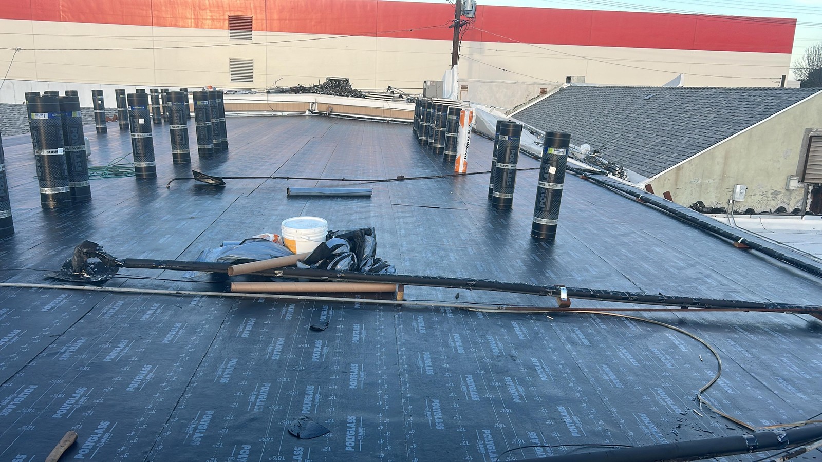 Rolls of black Polyglass roofing material standing upright on a flat low-slope roof beside torch hoses, a bucket and tarps
