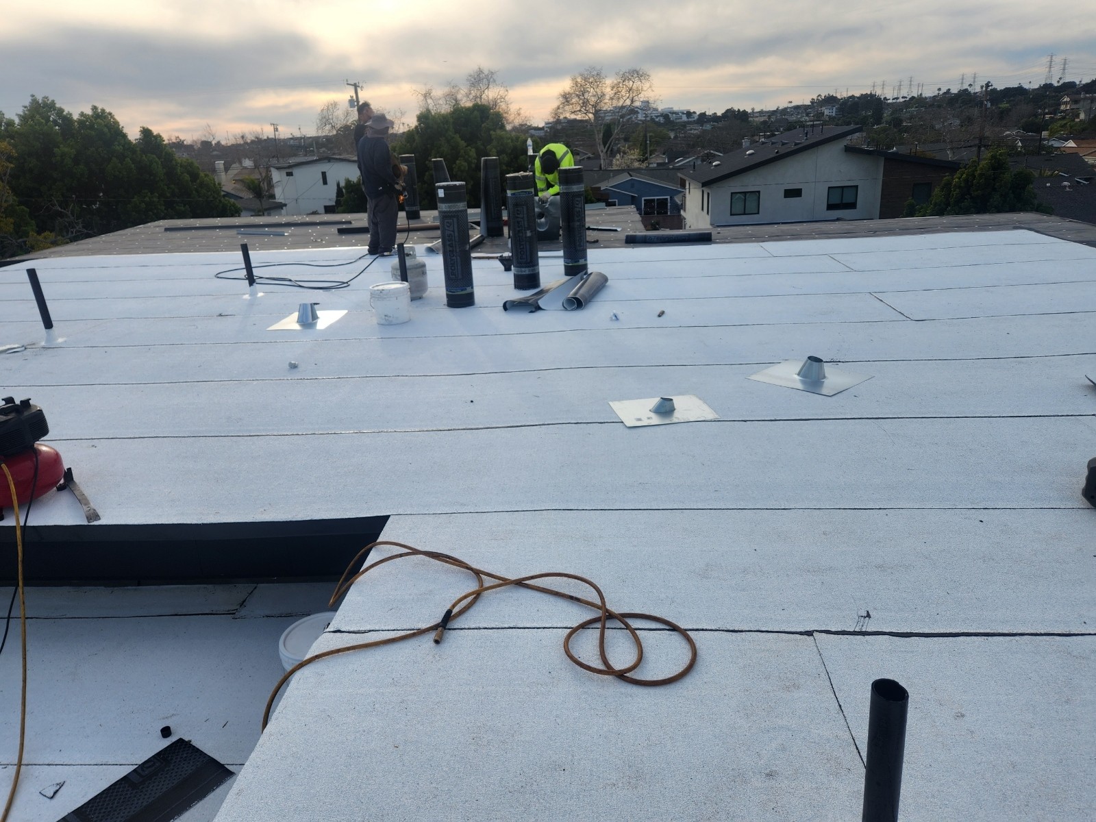 Two workers on a white flat roof with metal vent flashings, upright material rolls and a coiled hose.