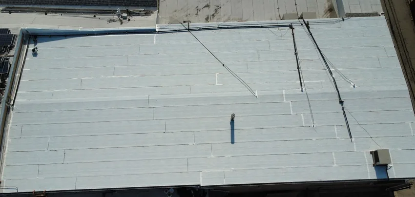 Overhead view of a pale low-slope roof with long parallel seams, cables, a small vent pipe and a rooftop unit
