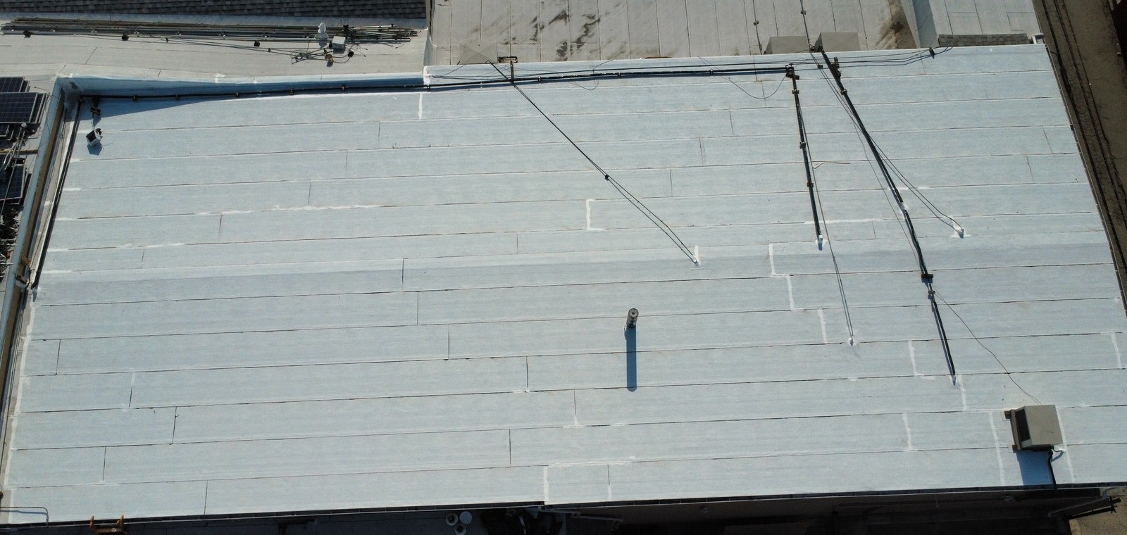 Overhead view of a pale low-slope roof with long parallel seams, cables, a small vent pipe and a rooftop unit