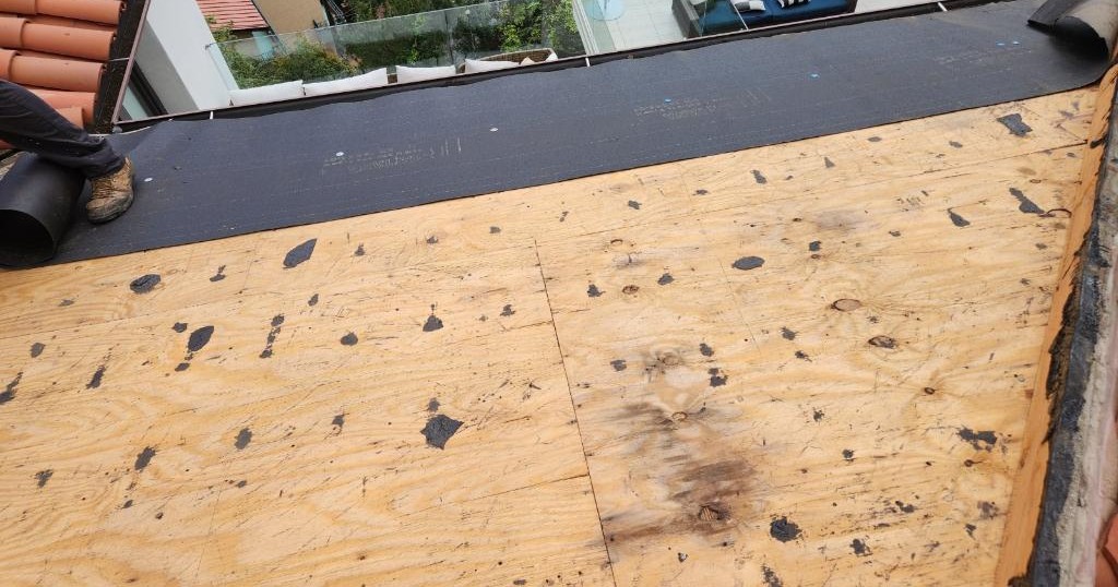 Stripped plywood roof deck with tar marks, clay tiles along the edges and a dark rolled sheet above.