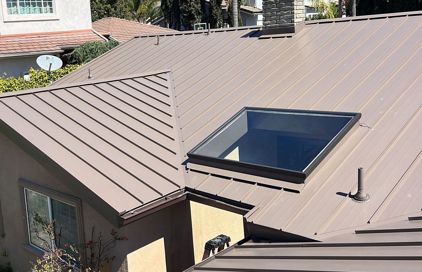 Brown standing-seam metal roof with a glass skylight, vent pipes and a stucco wall