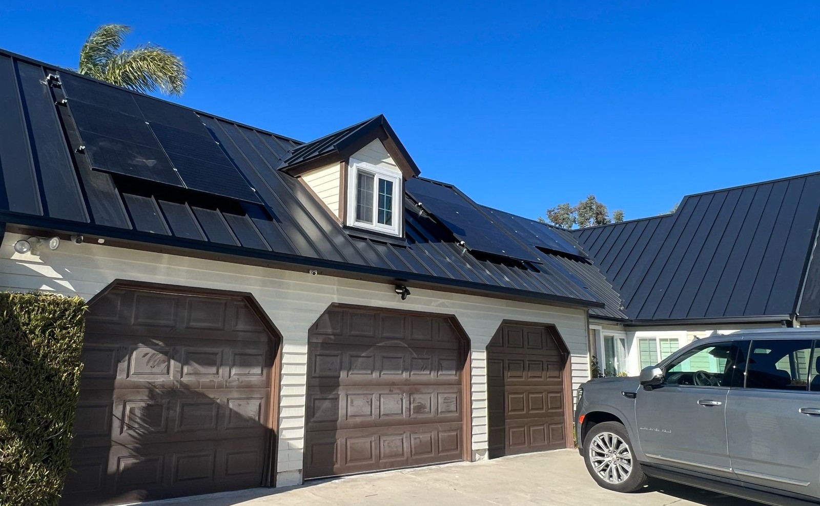 Black standing-seam metal roof with a dormer window and solar panels above three brown garage doors