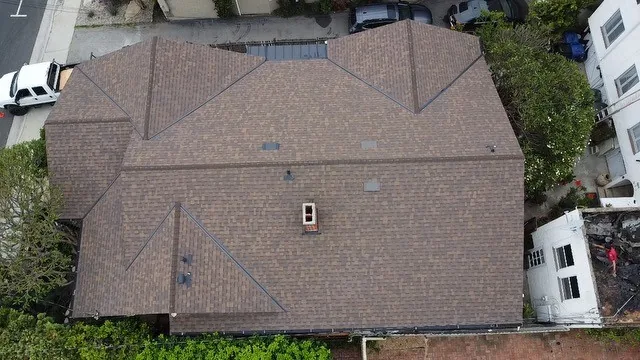 Aerial view of a brown asphalt shingle roof with multiple hips, valleys and a capped chimney