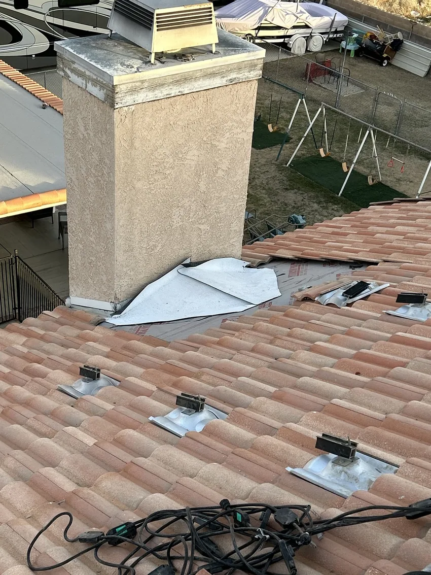 Clay tile roof with tiles lifted beside a stucco chimney, exposing underlayment and a white flashing sheet
