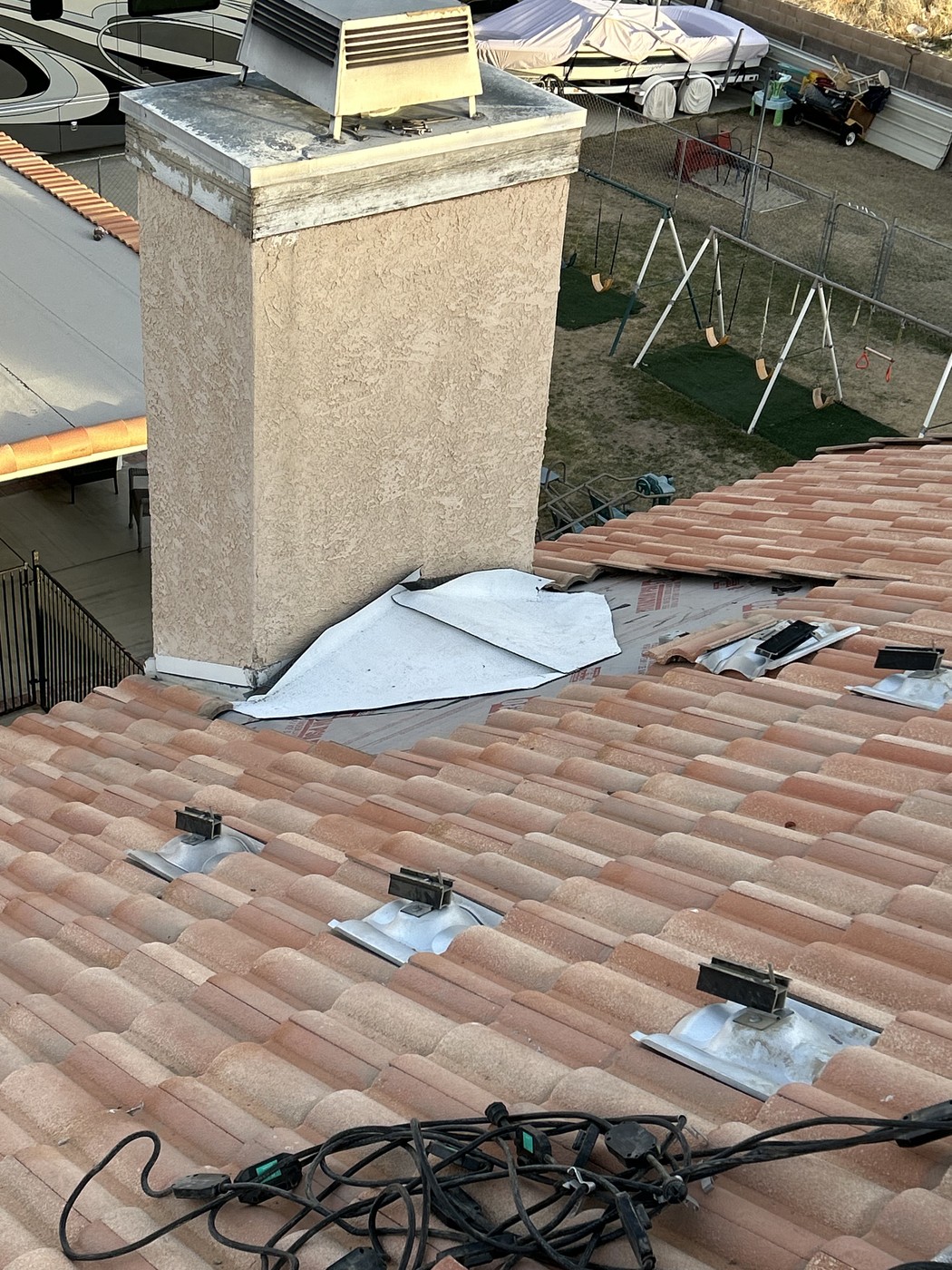 Terracotta barrel tiles lifted near a stucco chimney, with exposed underlayment, bolted metal brackets and a coiled black cable.