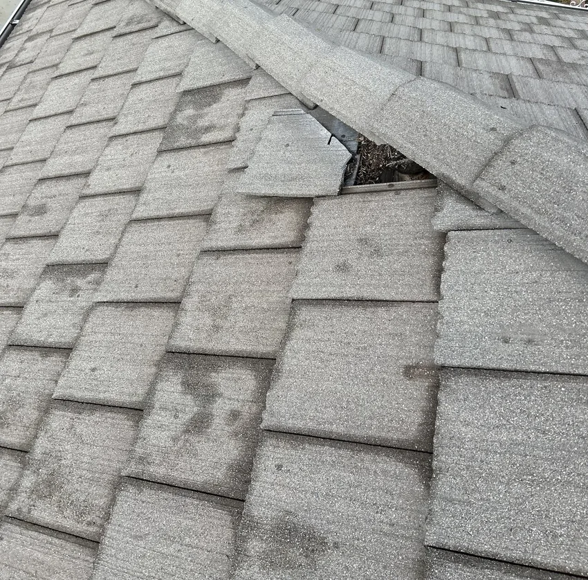 Gray granulated roof covering where hip cap pieces are missing, leaving a gap over metal flashing and dried leaf debris
