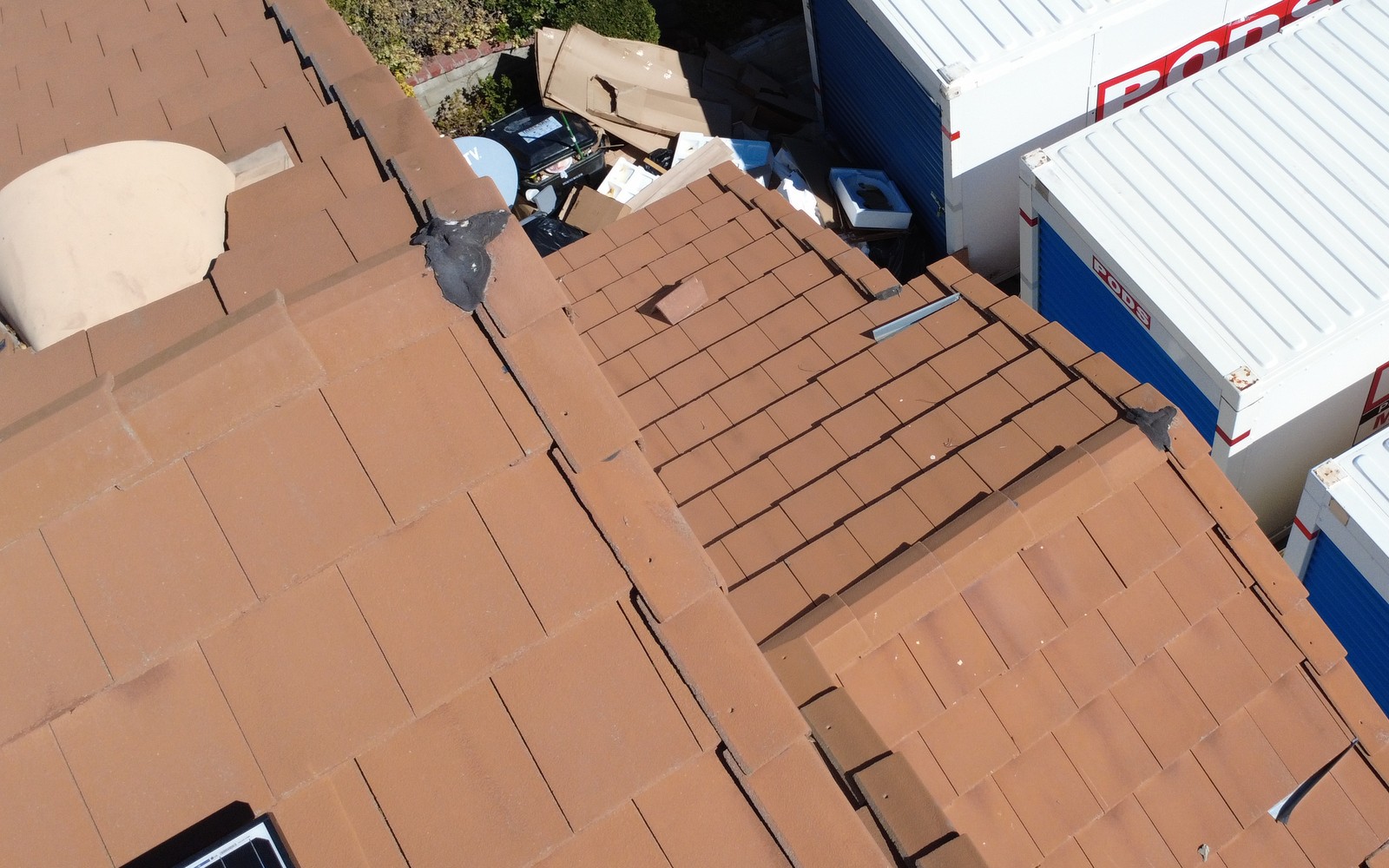 Overhead view of brown flat-profile tile roof slopes and ridge caps beside white and blue storage containers