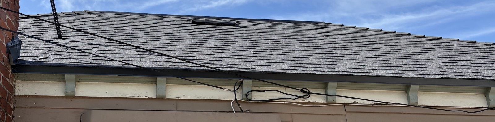 Gray asphalt shingle roof slope with a small vent, dark drip edge and fascia boards under overhead wires