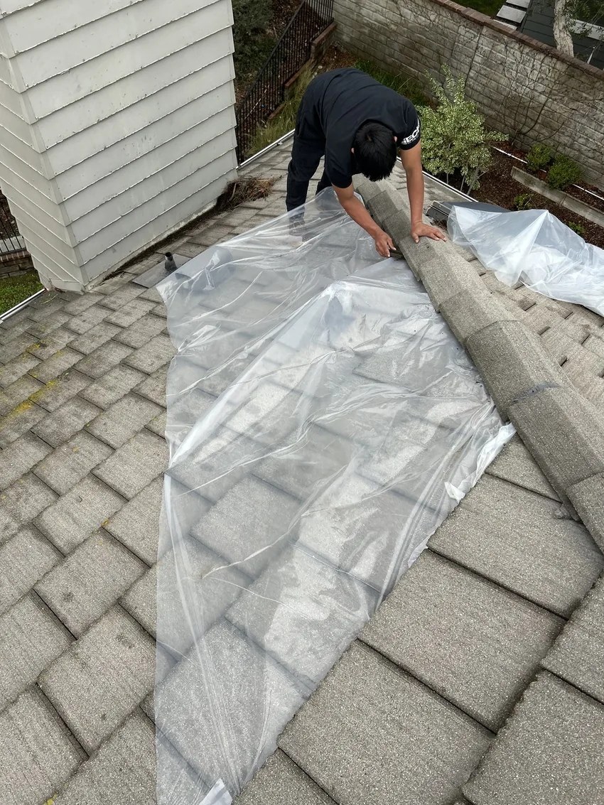 Worker in a dark shirt on a gray roof, handling a ridge piece above clear plastic sheeting