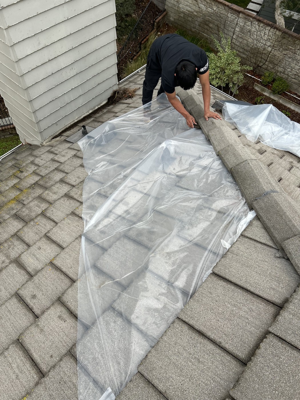 Worker in a dark shirt on a gray roof, handling a ridge piece above clear plastic sheeting