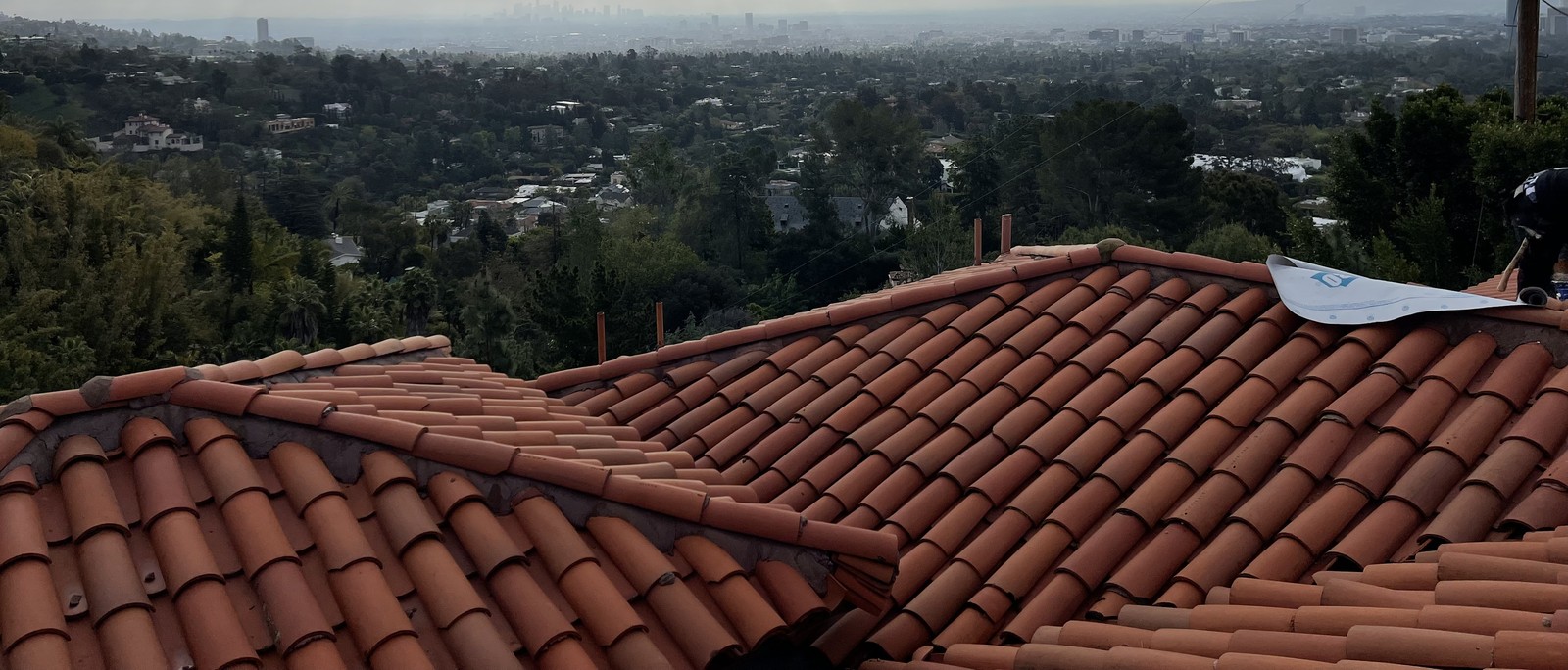 Terracotta barrel tile roof with hip ridges above a wooded hillside and a hazy city skyline