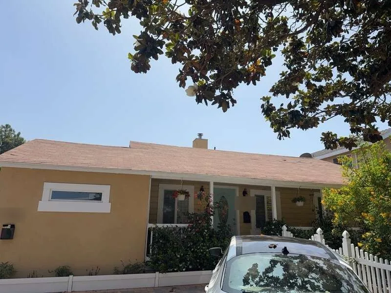 Single-story yellow stucco house with a light tan shingle roof, front porch and white picket fence
